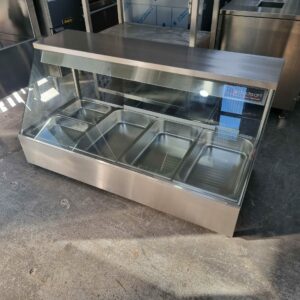 The WOODSON 4 MODULE BAIN MARIE, a stainless steel display case with a glass front and five metal pans, sits on a concrete floor—ideal commercial catering equipment for any industrial or commercial kitchen. MRCE