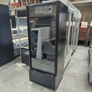 A SKOPE SINGLE DOOR DISPLAY FRIDGE from Melbourne Catering Equipment stands in a warehouse, its black frame and glass door showing empty shelves inside, with other commercial appliances and stacked chairs visible nearby. MRCE