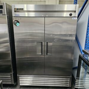 The TRUE REFRIGERATION 2 DOOR STORAGE FRIDGE, a large stainless steel unit with sturdy handles, is displayed at Melbourne Catering Equipment beside blue metal shelving and other commercial catering gear. MRCE