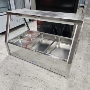 A stainless steel food display case with a glass front and three empty metal food trays, standing on a concrete floor in a commercial setting. MRCE