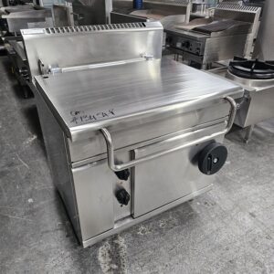 A large stainless steel commercial kitchen appliance with a flat lid, front handle, control knobs, and storage below, placed on a concrete floor among other industrial kitchen equipment. MRCE