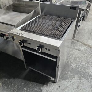 A commercial stainless steel char grill with a grated cooking surface, control knobs, and an open shelf below, placed on a concrete floor among other kitchen equipment. MRCE