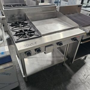 A commercial stainless steel kitchen range with two gas burners, a flat griddle top, control knobs in front, and an open lower shelf, situated in an industrial kitchen setting. MRCE