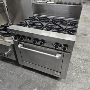 A commercial stainless steel gas range with six burners and an oven, situated in an industrial kitchen setting with a concrete floor. MRCE