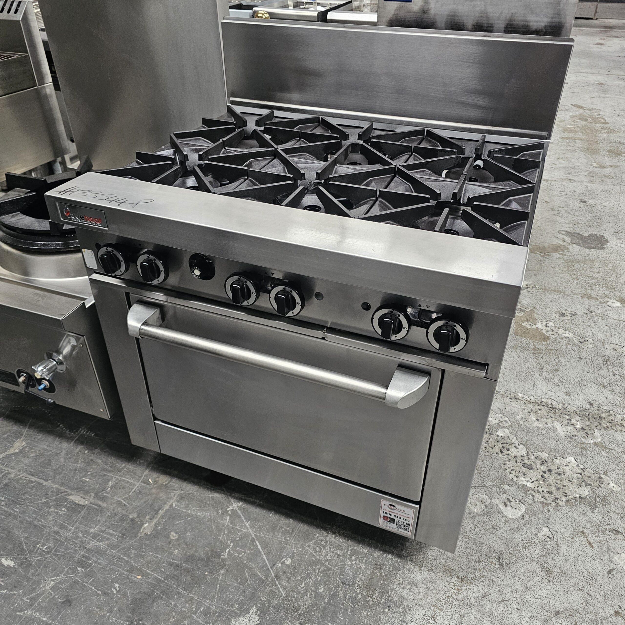 A commercial stainless steel gas range with six burners and an oven, situated in an industrial kitchen setting with a concrete floor. MRCE