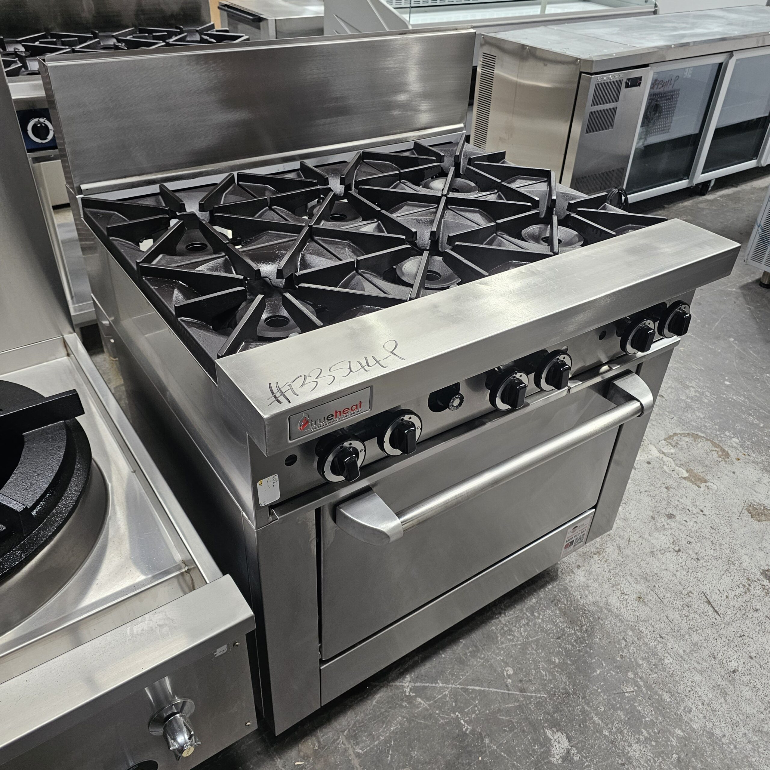 A commercial stainless steel gas range with six burners and an oven below, standing in a kitchen equipment showroom. The range has control knobs and is labeled with a tag reading #335448. MRCE
