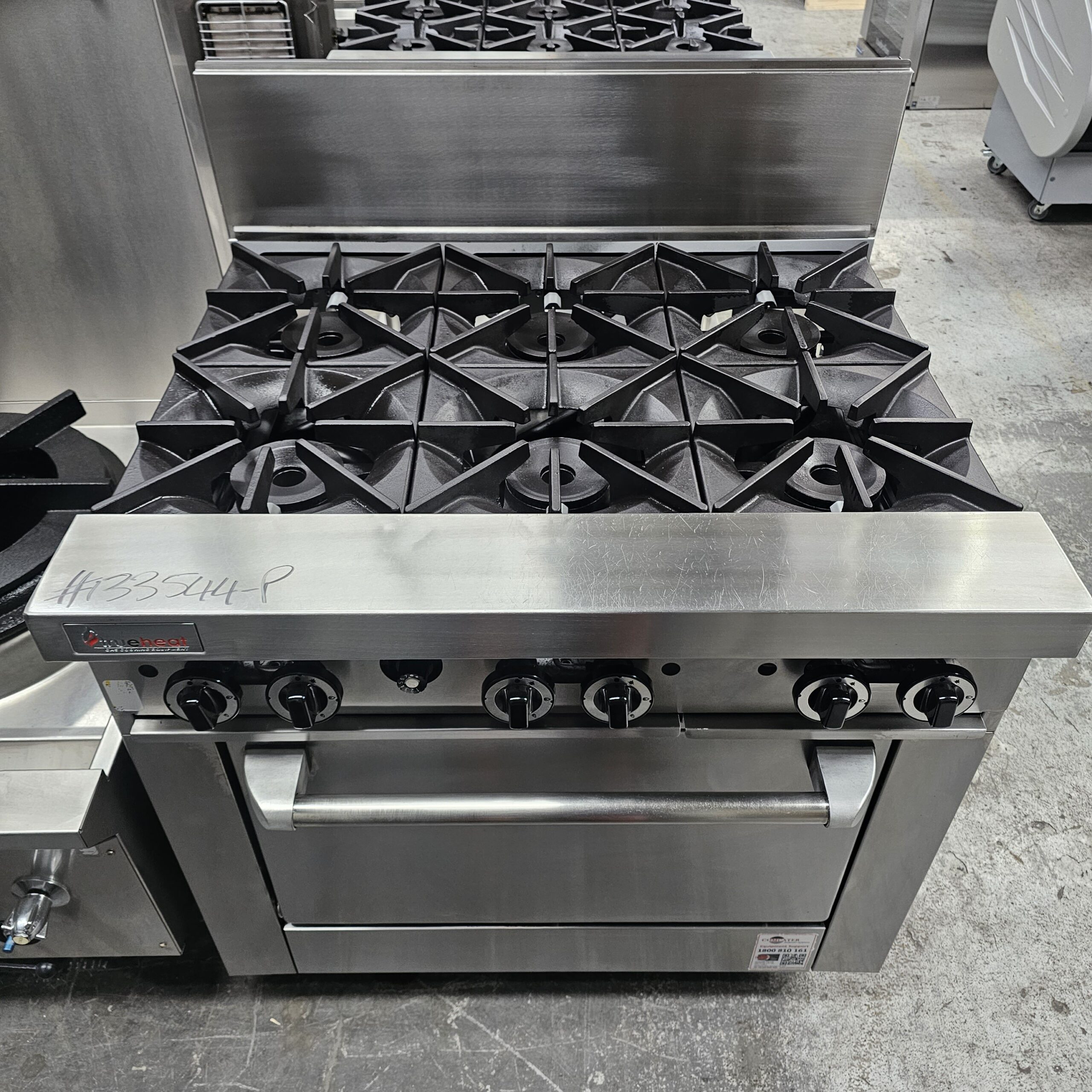 A commercial stainless steel gas range with six burners and an oven below. The appliance has black control knobs and is situated in an industrial kitchen setting. MRCE