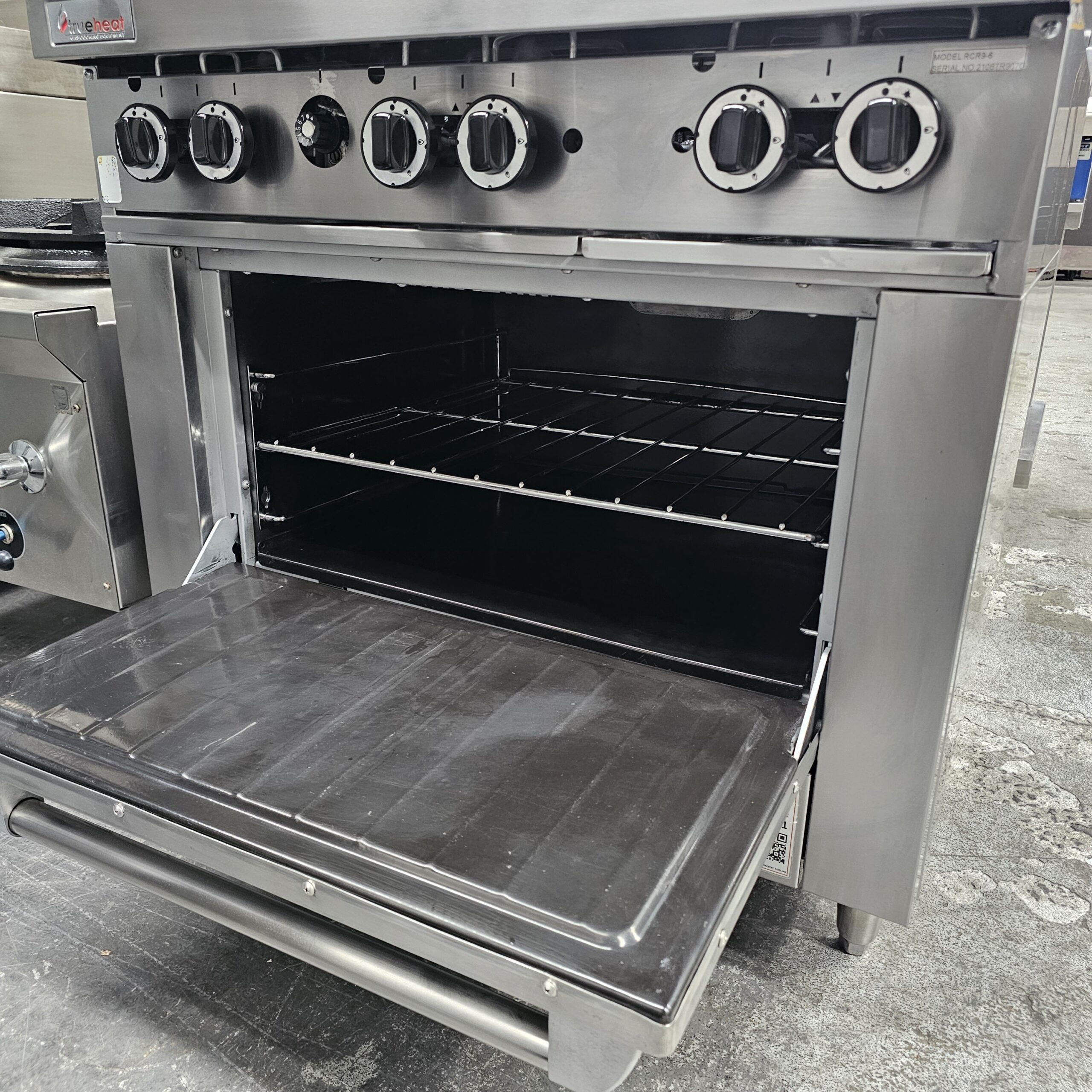 A stainless steel commercial oven with its door open, showing two wire racks inside. Control knobs are visible on the upper front panel. The oven is set in an industrial kitchen environment. MRCE