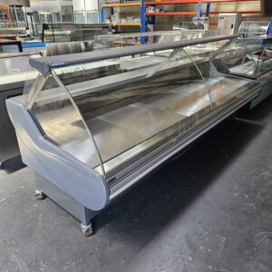 A large, empty refrigerated display case with curved glass front and metal shelves sits on a dark floor in a commercial space, surrounded by other equipment. MRCE