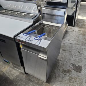 A commercial deep fryer with two metal baskets and blue handles is shown in a stainless steel kitchen setting. The price $1550.00 is written on the fryer’s surface. The surrounding area has other kitchen equipment. MRCE