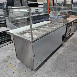 A stainless steel commercial food display counter with a glass sneeze guard, featuring six metal food trays. The unit is situated in an industrial kitchen or showroom setting. MRCE