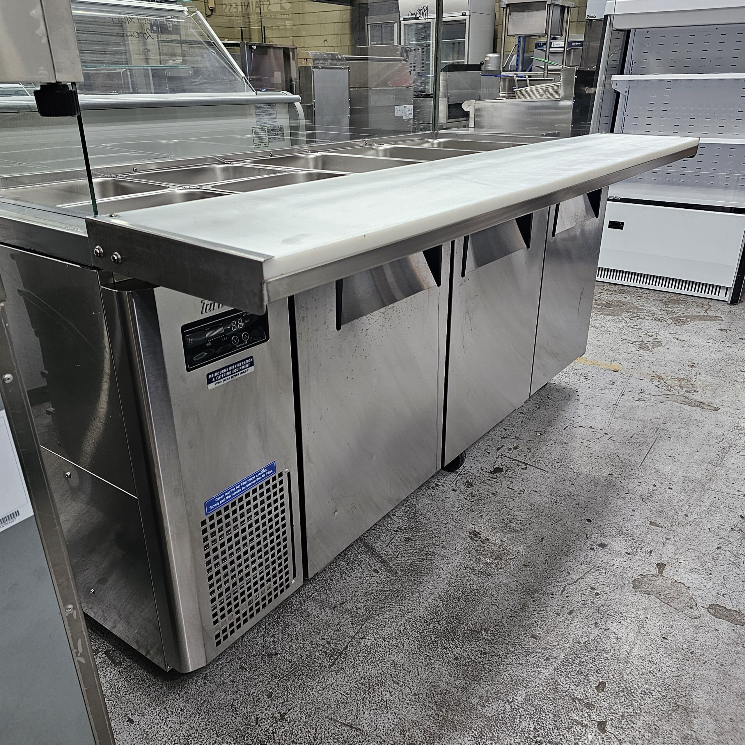 A stainless steel commercial prep table with refrigerated storage and multiple metal food trays on top, standing on a textured floor in a commercial kitchen or food prep area. MRCE