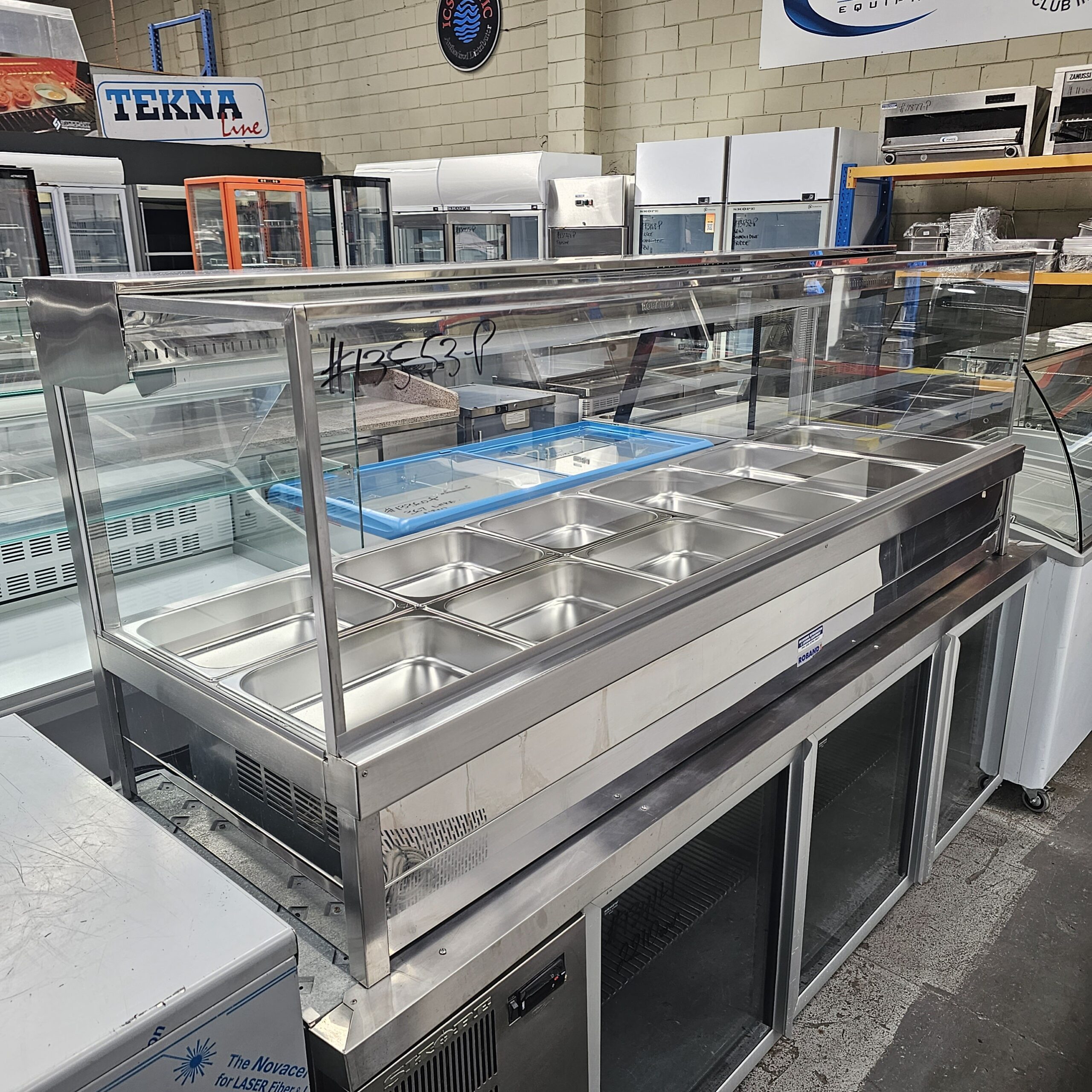 A stainless steel refrigerated food display case with multiple empty metal trays, covered by a glass sneeze guard, is situated in an appliance showroom with various refrigerators and freezers in the background. MRCE