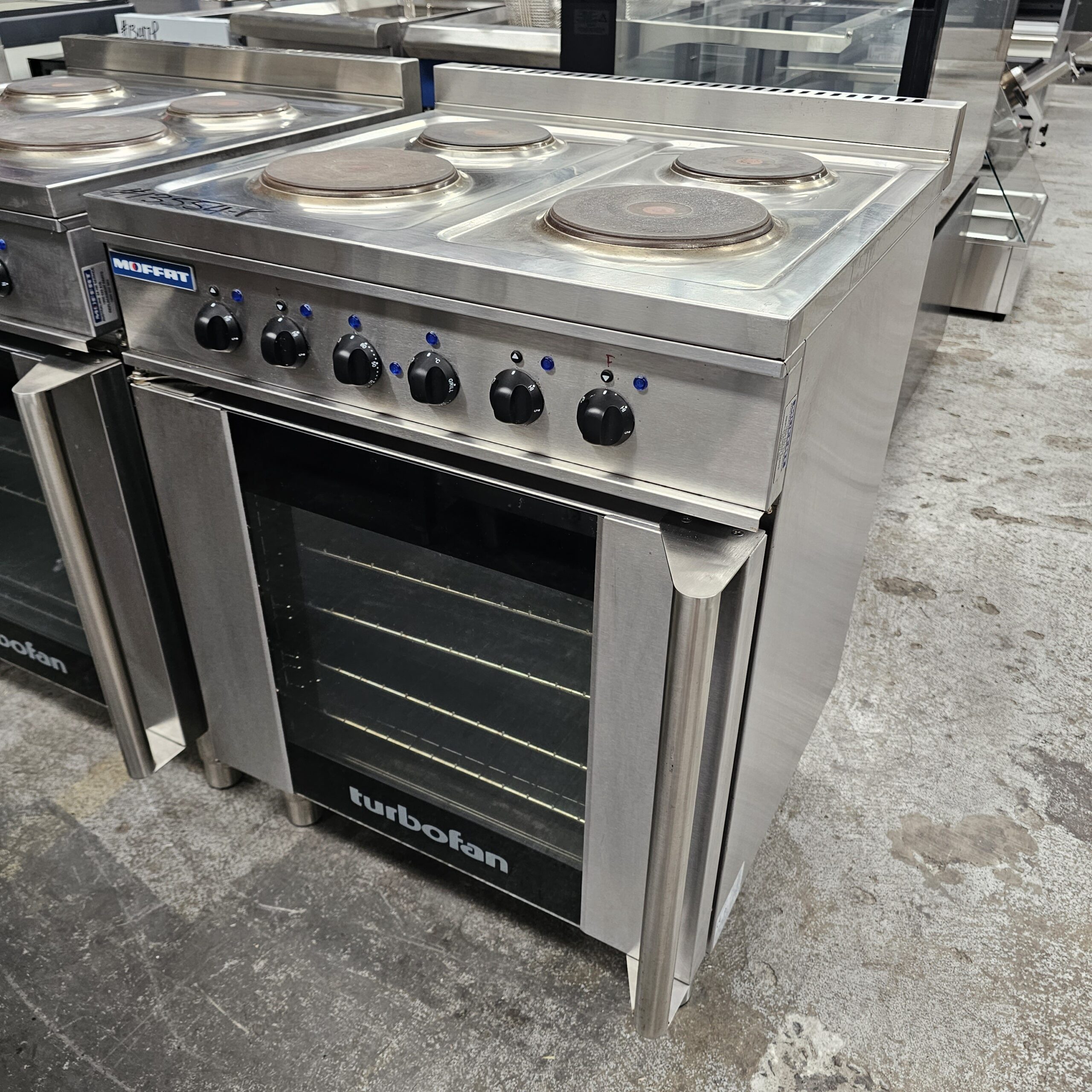 A stainless steel electric stove with four burners on top, control knobs on the front, and a glass-fronted oven door labeled turbofan, standing on a concrete floor in a commercial kitchen setting. MRCE