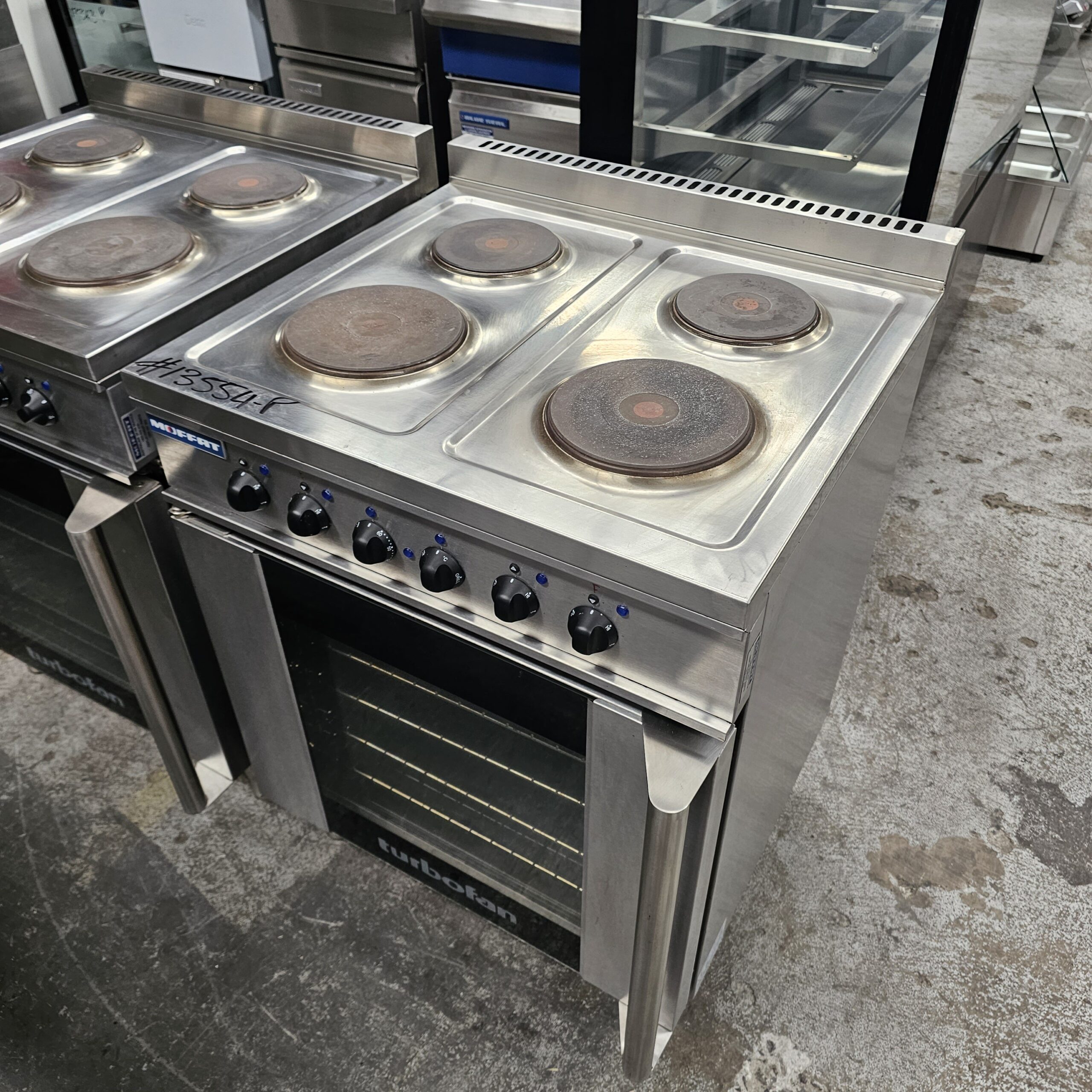 A stainless steel electric stove with four round burners and a black oven door, situated on a concrete floor in what appears to be a commercial kitchen or appliance showroom. MRCE