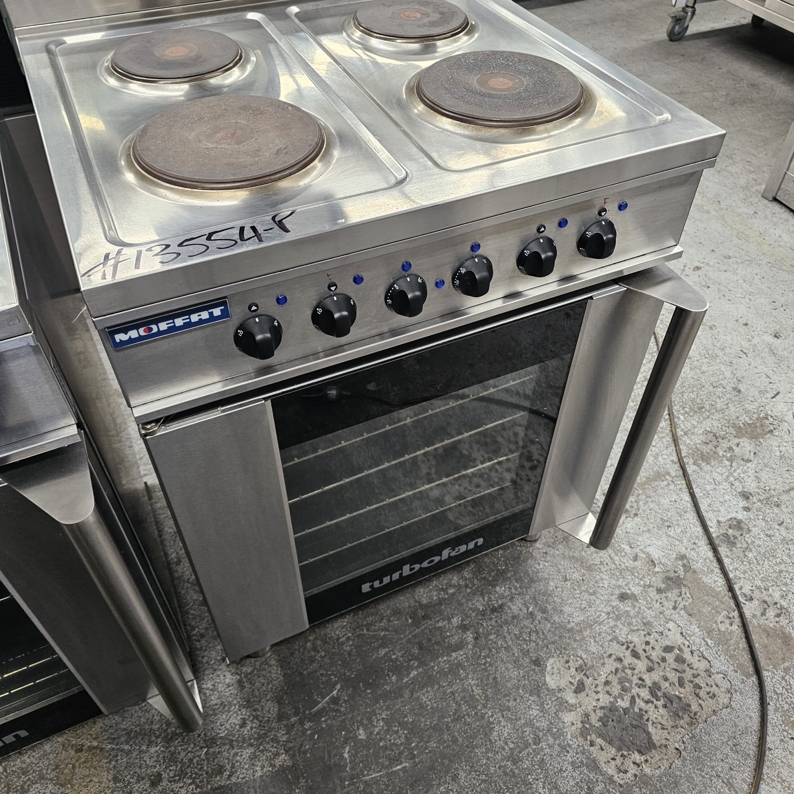 A stainless steel Moffat electric stove with four coil burners, six black control knobs, and a glass-fronted oven labeled turbofan, sitting on a concrete floor in an industrial setting. MRCE