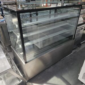 A large, empty glass display case with metal shelves and a stainless steel base is positioned on a concrete floor in a commercial kitchen or store setting. MRCE
