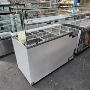 A commercial food display unit with a glass sneeze guard, featuring multiple stainless steel compartments for food storage, stands on a concrete floor in a warehouse setting. Handwritten prices are visible on the glass. MRCE