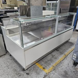 A large, empty refrigerated display case with glass sides and a metal interior, situated in what appears to be a commercial kitchen or warehouse setting. MRCE
