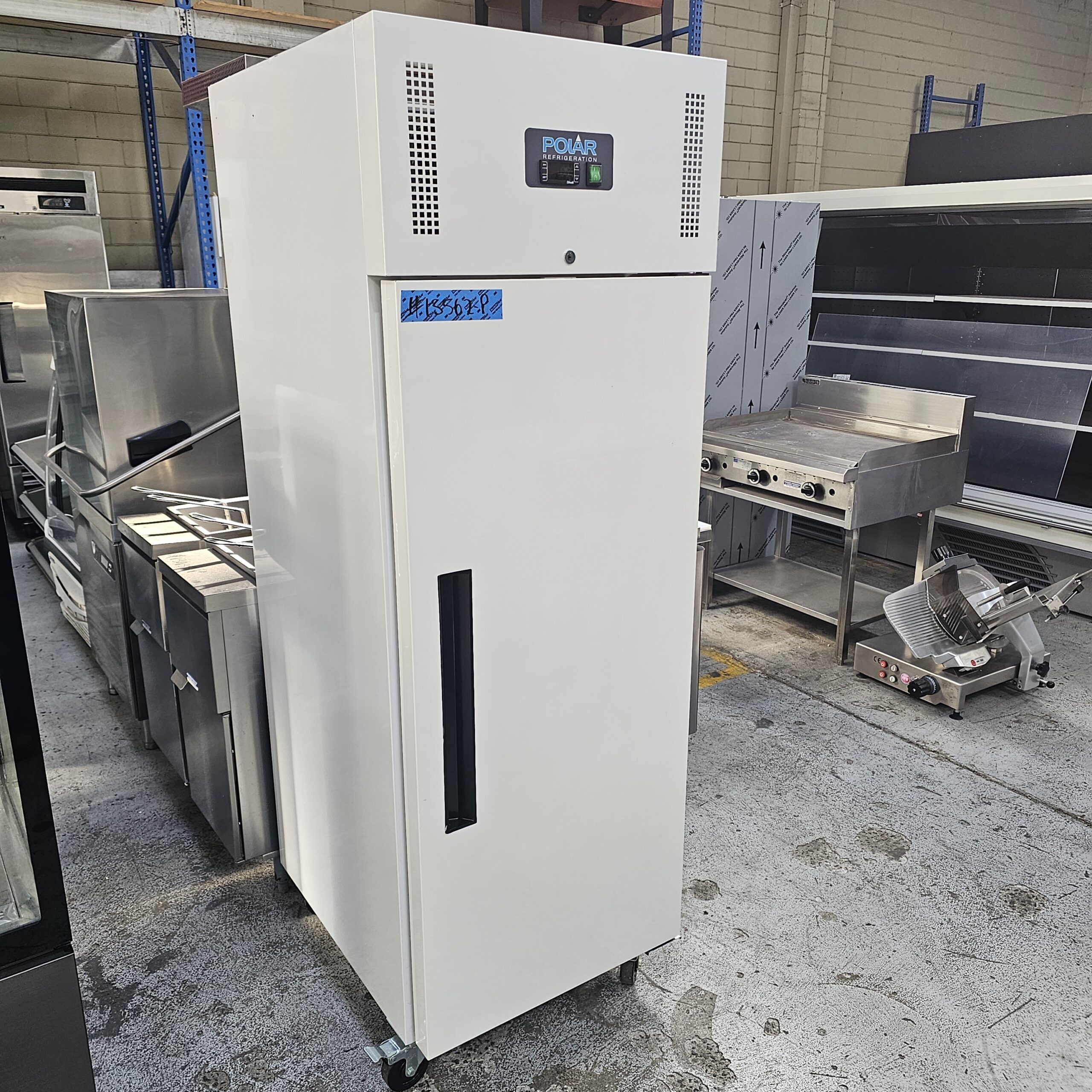 A tall, white commercial refrigerator with a digital display and air vents on top stands in a warehouse. It is surrounded by stainless steel kitchen equipment and has wheels for mobility. MRCE