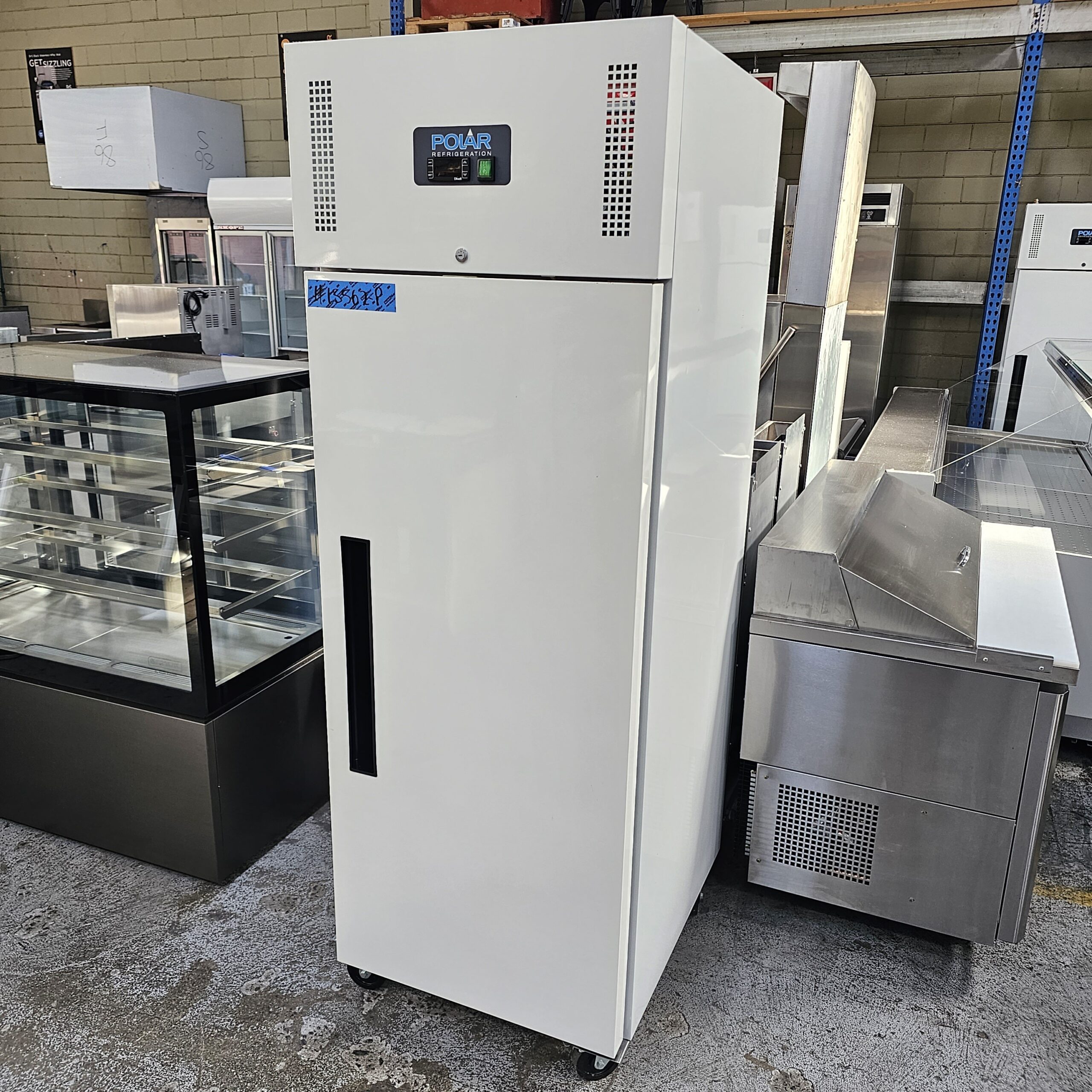 A tall, white commercial refrigerator on wheels stands in a warehouse with other stainless steel kitchen equipment and appliances in the background. The refrigerator has a digital display and a vertical handle. MRCE