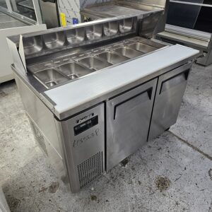 A stainless steel commercial prep fridge with two doors and a top section holding multiple ingredient trays, displayed in a showroom on a concrete floor. A price of £1553.29 is marked on the front. MRCE