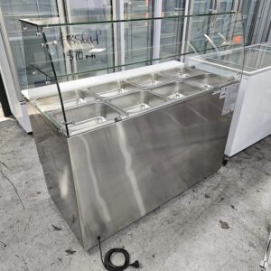 A stainless steel commercial food display counter with eight covered compartments, a glass sneeze guard, and a power cord, situated indoors on a concrete floor. MRCE