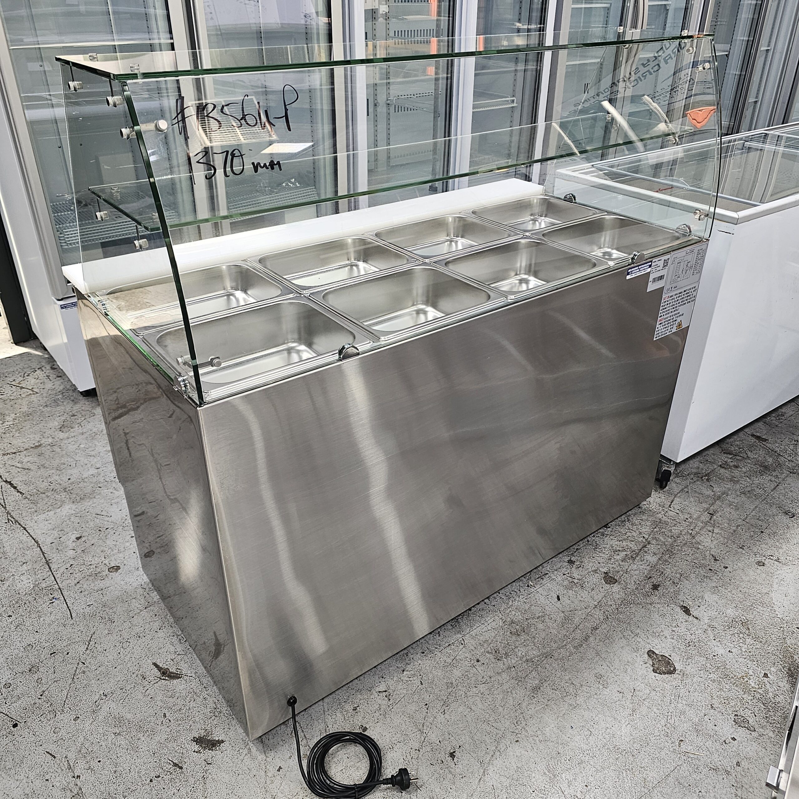 A stainless steel commercial food display counter with eight covered compartments, a glass sneeze guard, and a power cord, situated indoors on a concrete floor. MRCE