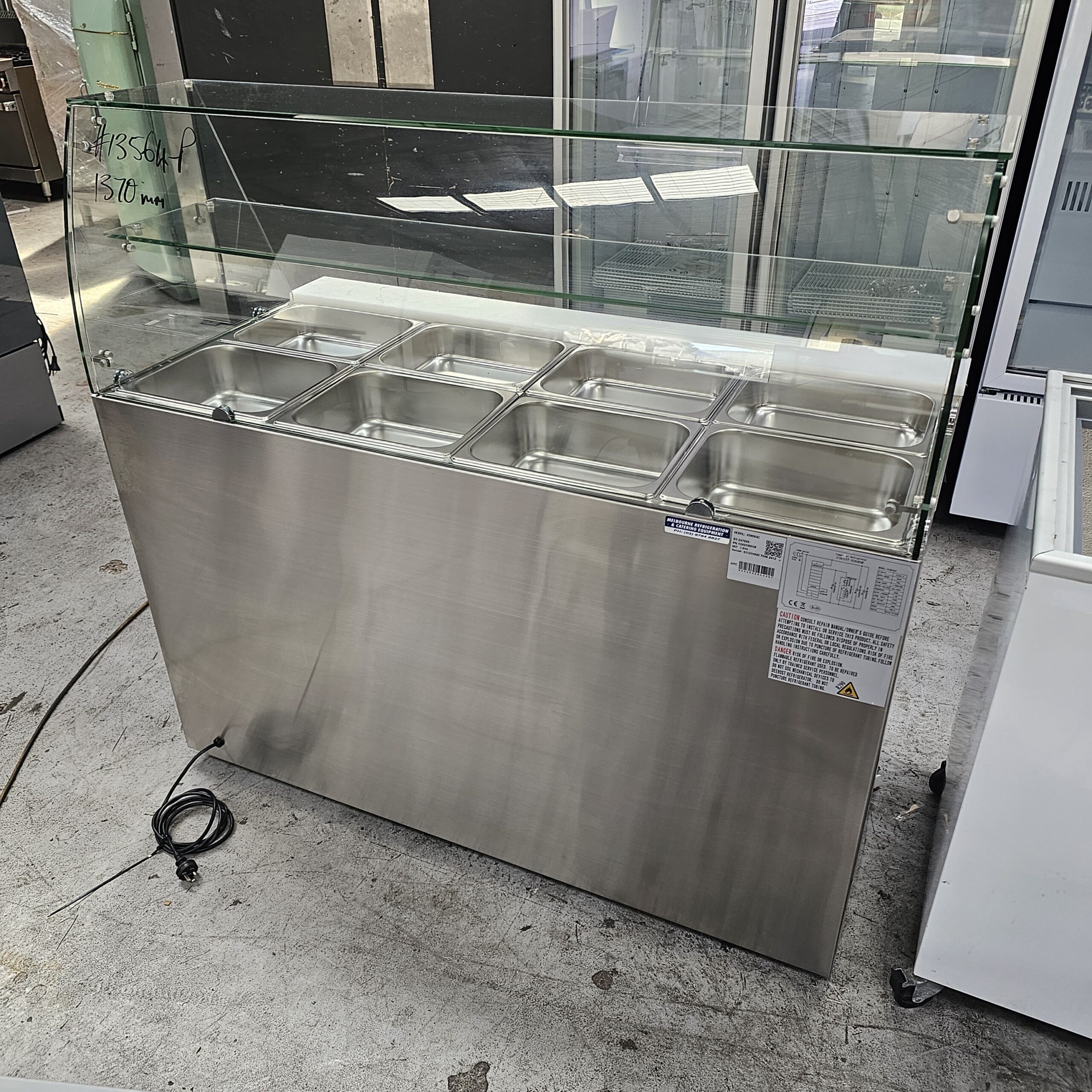 A stainless steel commercial food display counter with eight metal food trays and a glass sneeze guard, set on a concrete floor in an industrial space. An electrical cord is on the ground beside it. MRCE