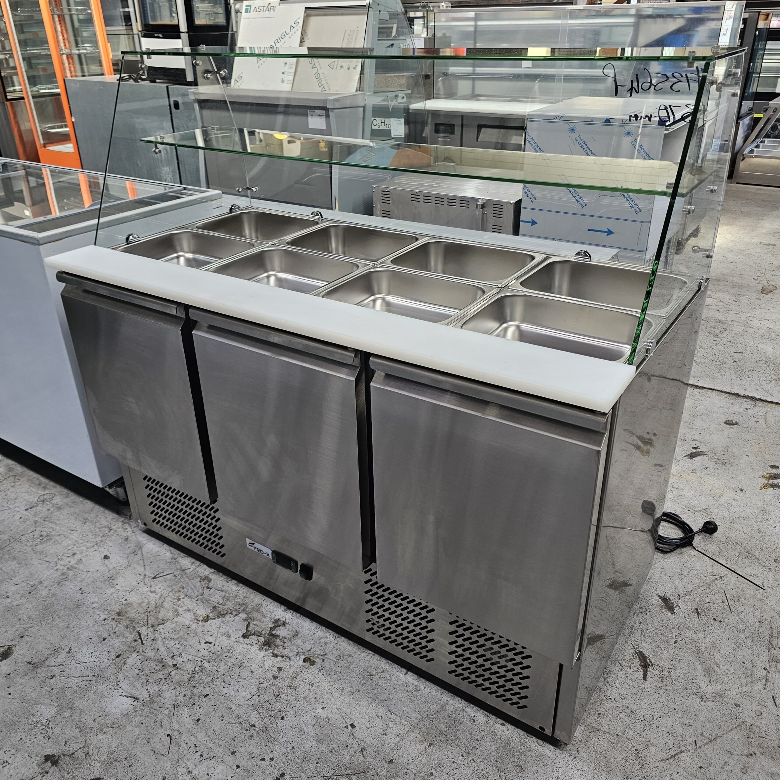 A stainless steel refrigerated food prep station with three doors below and six metal food pans on top, covered by a glass sneeze guard, is seen in a commercial kitchen setting. MRCE
