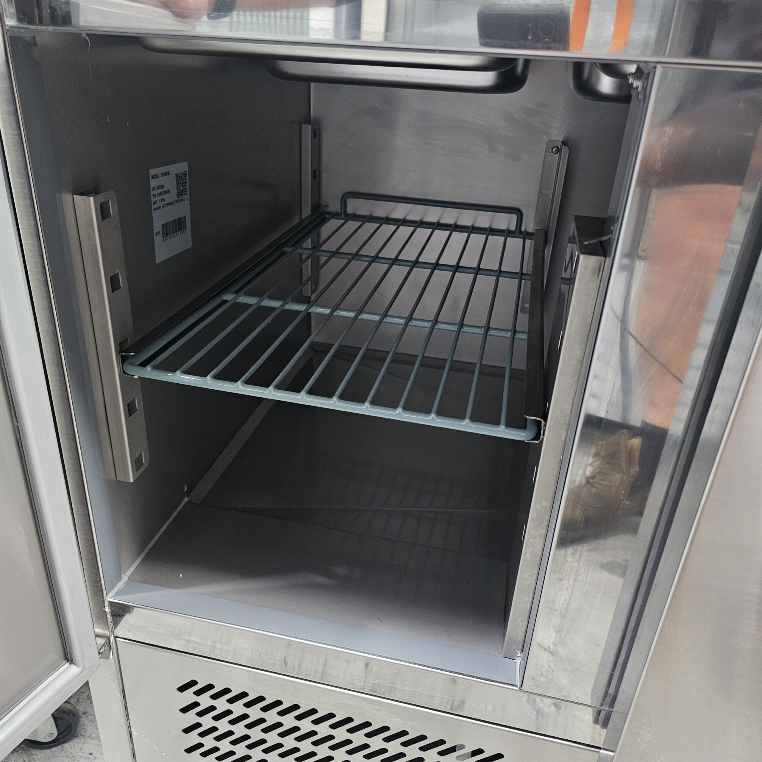 A close-up view of an open, empty stainless steel commercial refrigerator with a single wire shelf inside and a vent at the bottom. MRCE