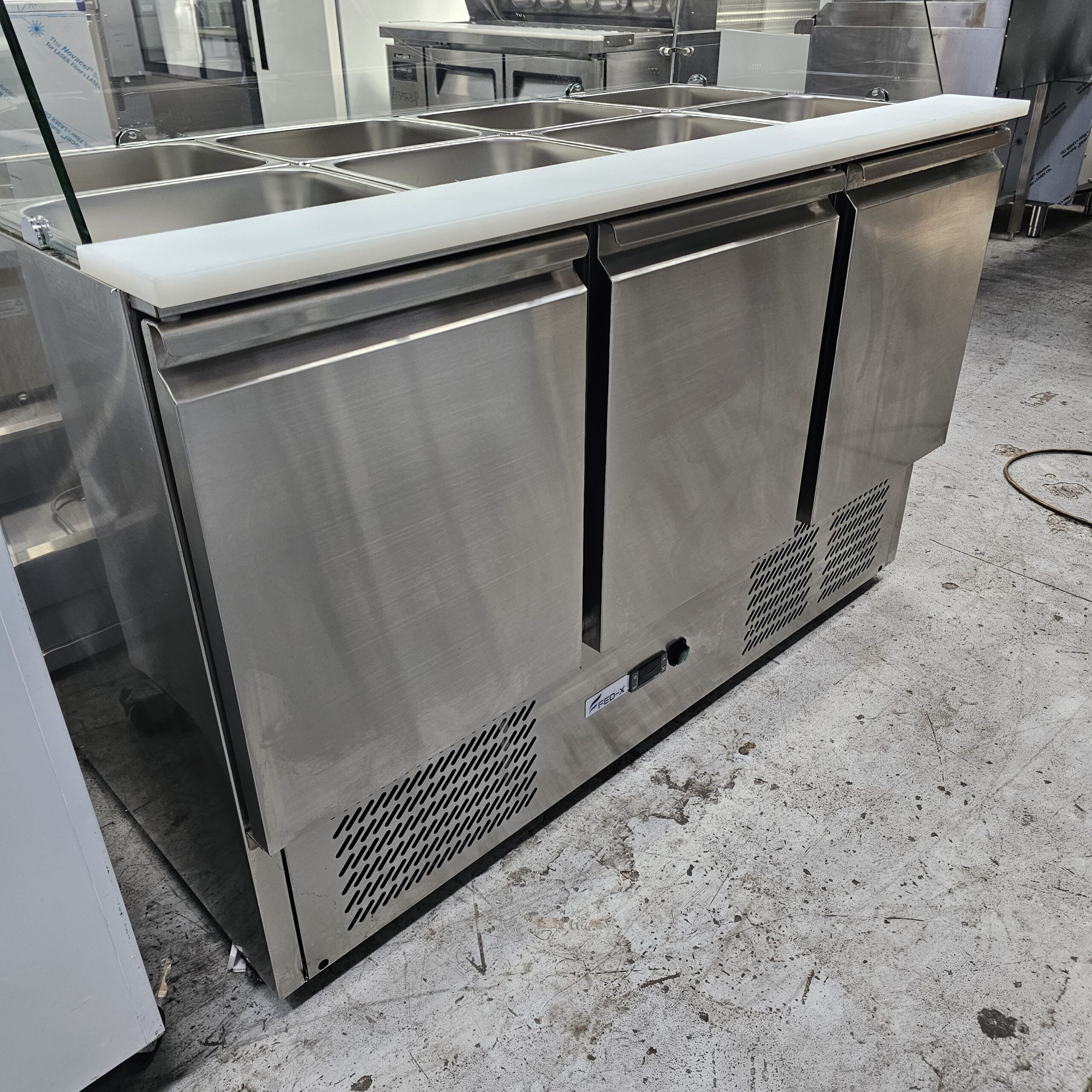 A stainless steel commercial refrigerator with three doors and a white countertop, featuring multiple food storage compartments on top, situated on a concrete floor in an industrial kitchen setting. MRCE