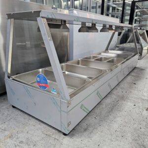A large, stainless steel commercial food buffet station with multiple empty food trays and overhead heat lamps, situated in an industrial or kitchen setting. MRCE