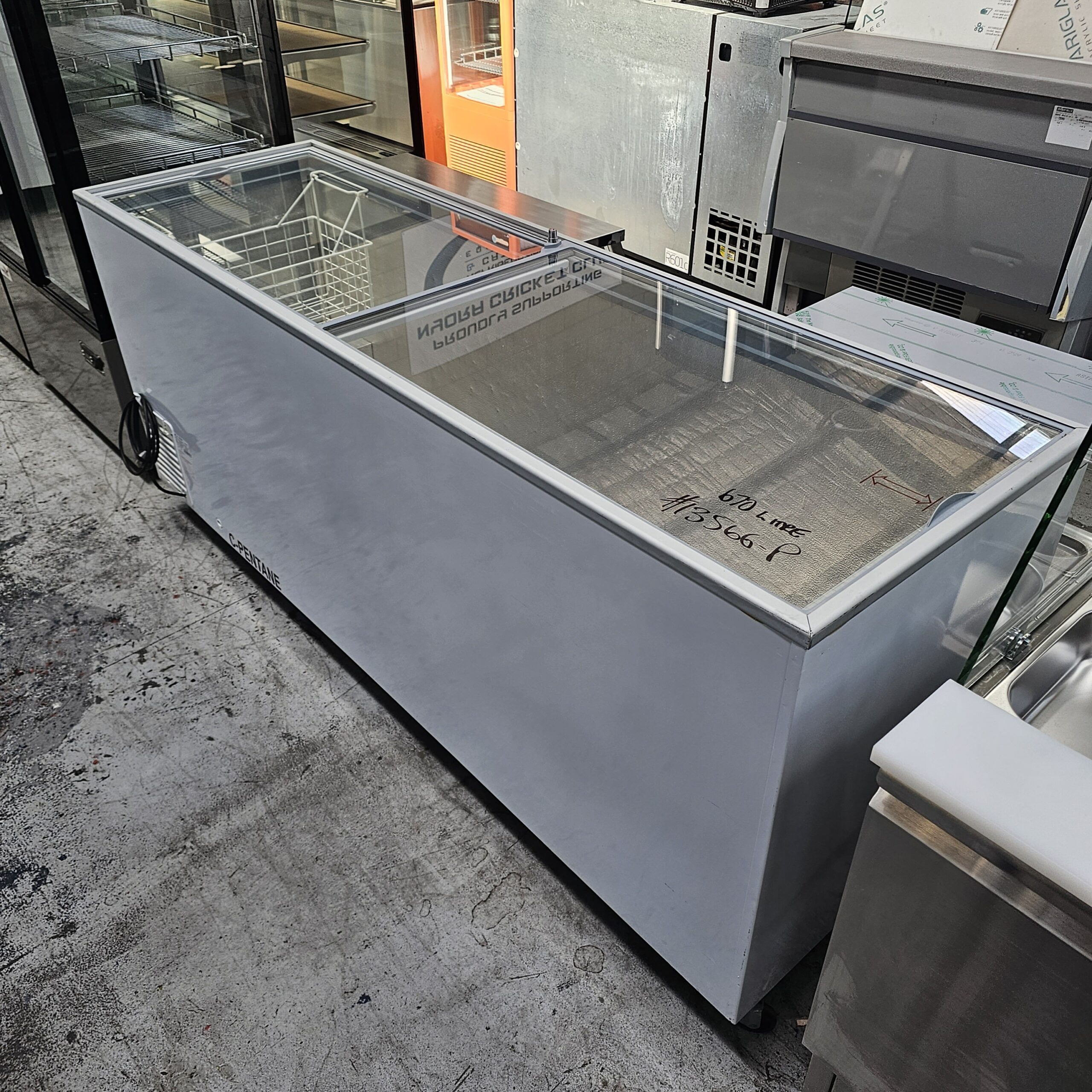 A large, rectangular commercial freezer with a sliding glass top is displayed in a showroom. The freezer is white with a silver frame and placed on a concrete floor among other appliances. MRCE