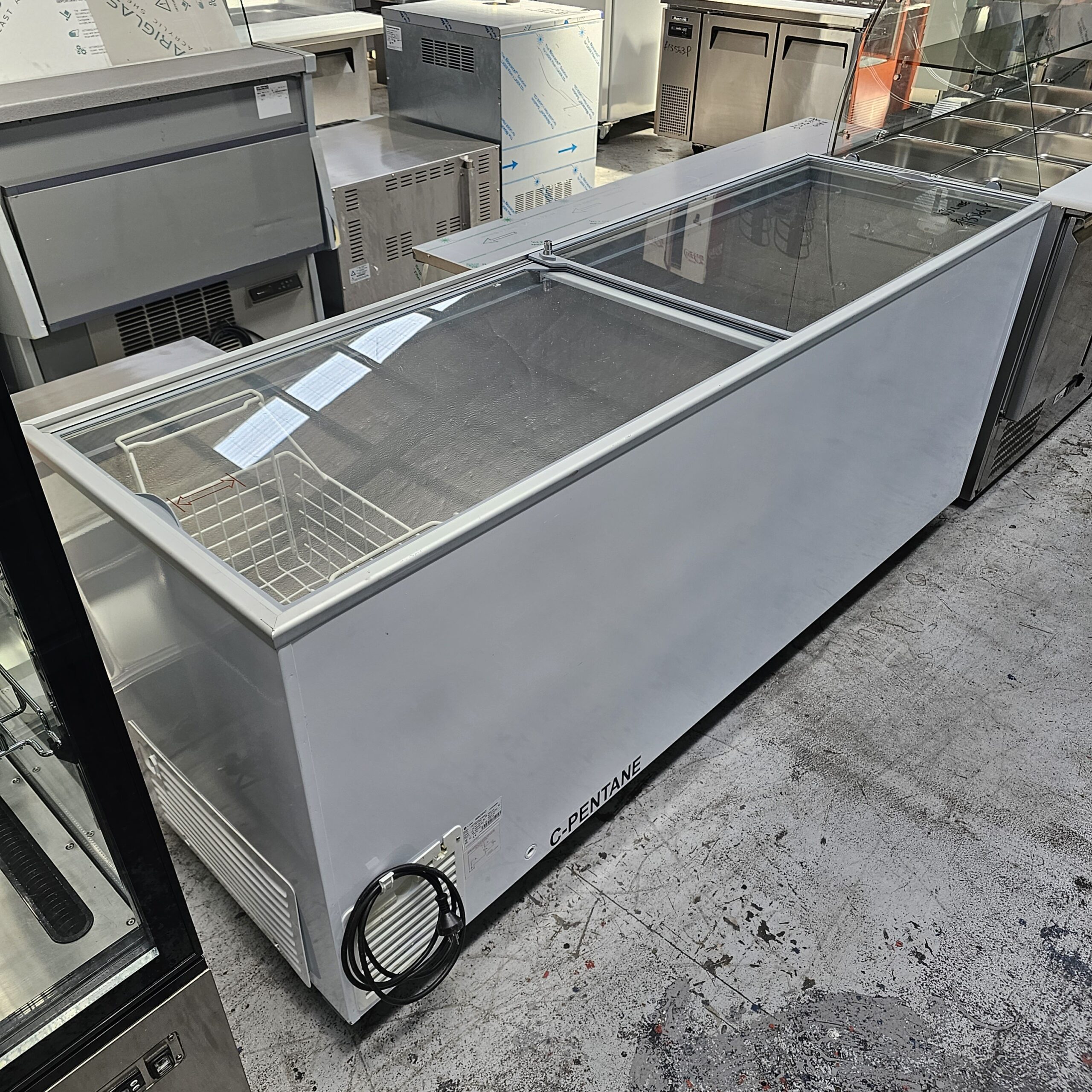 A large, white commercial chest freezer with a glass sliding lid and internal wire basket, placed on a concrete floor among other industrial kitchen appliances. MRCE