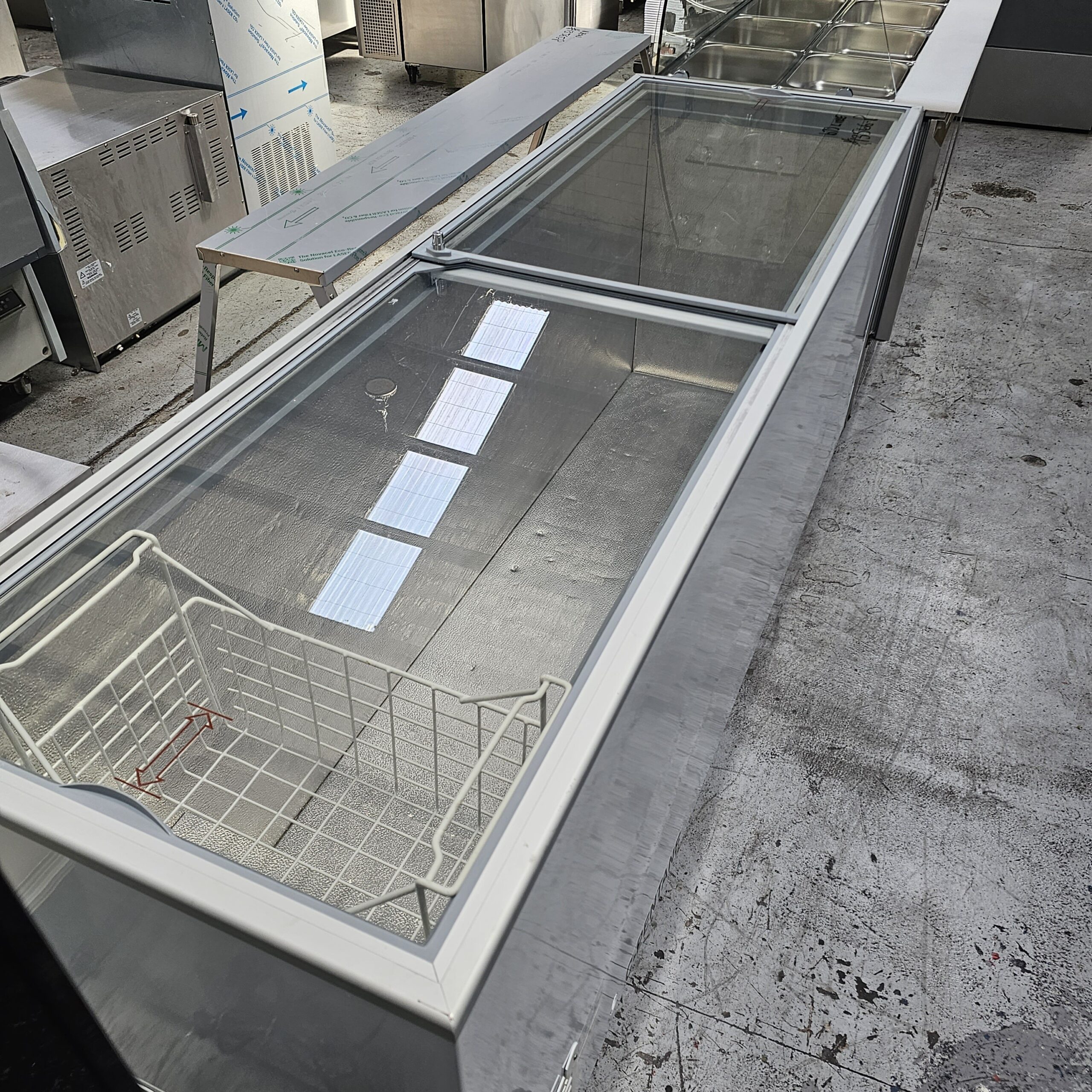A large, empty commercial freezer with a glass sliding lid and a wire basket inside is placed in an industrial kitchen with other stainless steel appliances and equipment nearby. MRCE
