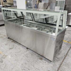 A stainless steel refrigerated food display case with a glass sneeze guard, featuring multiple metal food trays inside, placed on a concrete floor in an industrial setting. MRCE