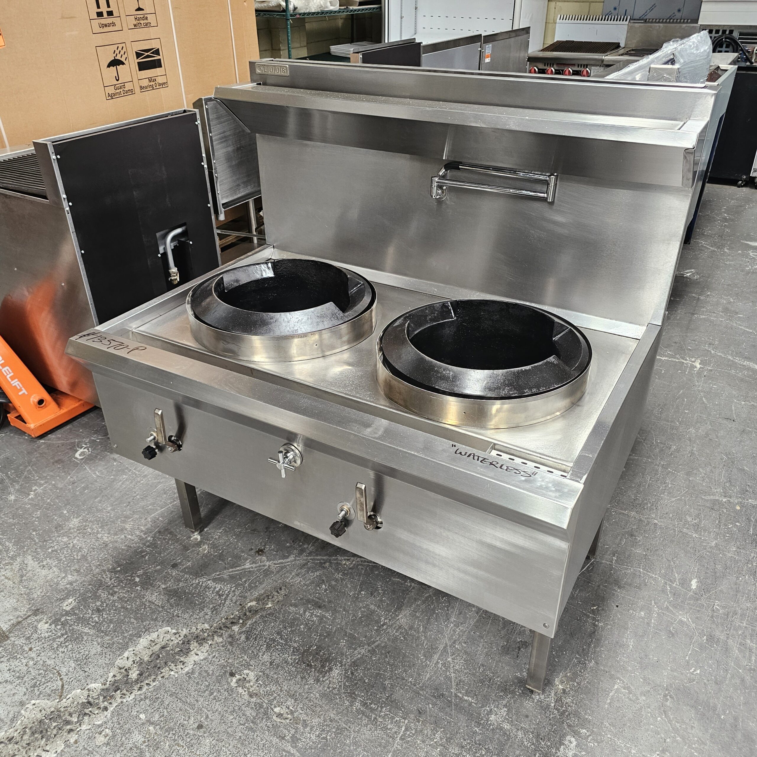 A commercial stainless steel double-burner wok range with two large round burners, side controls, and a backsplash, situated on a warehouse floor. MRCE