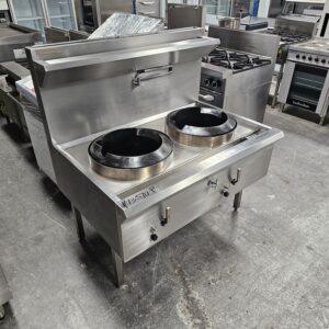 A stainless steel commercial double wok range with two large burners and control knobs, standing in an industrial kitchen equipment showroom. Other kitchen appliances are visible in the background. MRCE