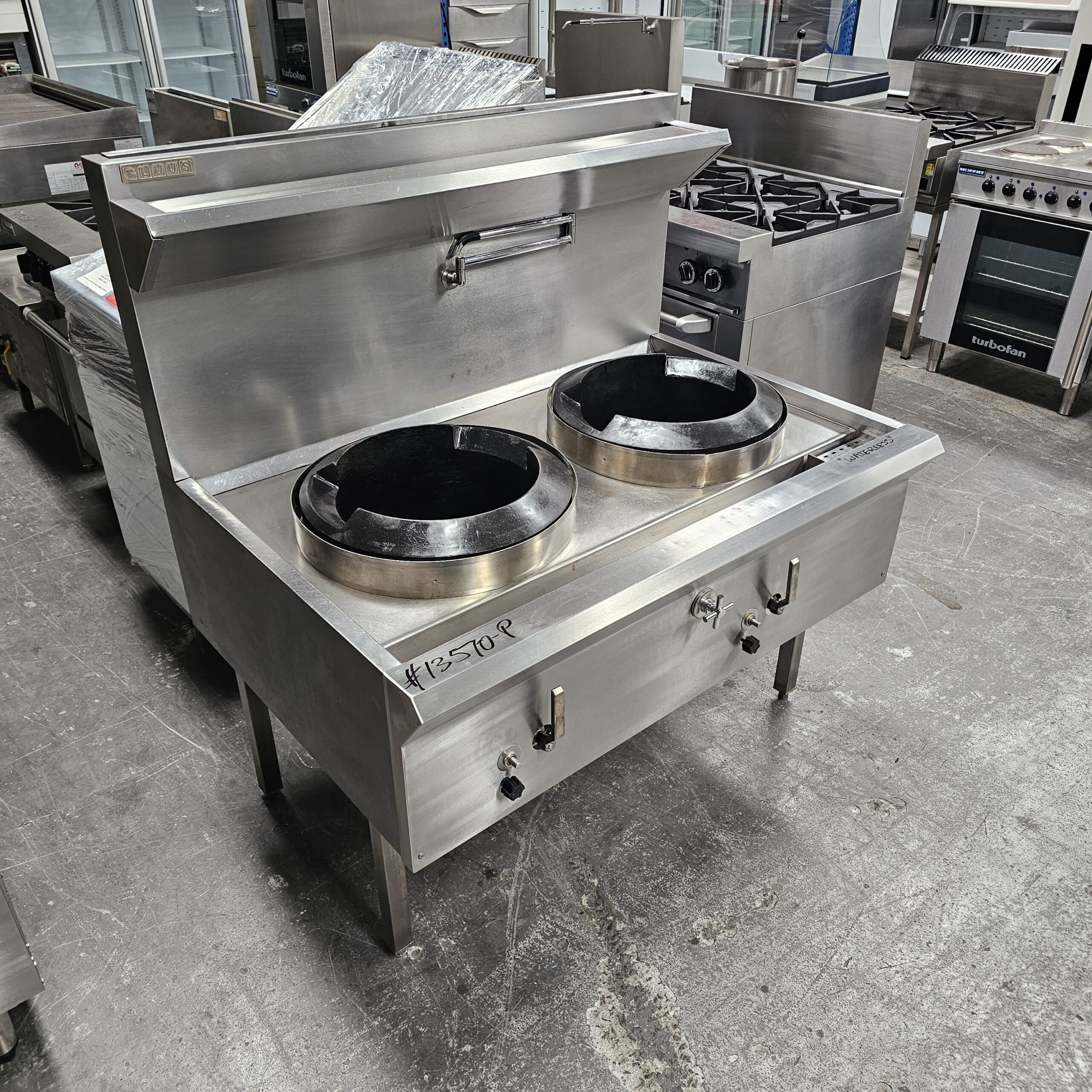 A stainless steel commercial double wok range with two large burners and control knobs, standing in an industrial kitchen equipment showroom. Other kitchen appliances are visible in the background. MRCE