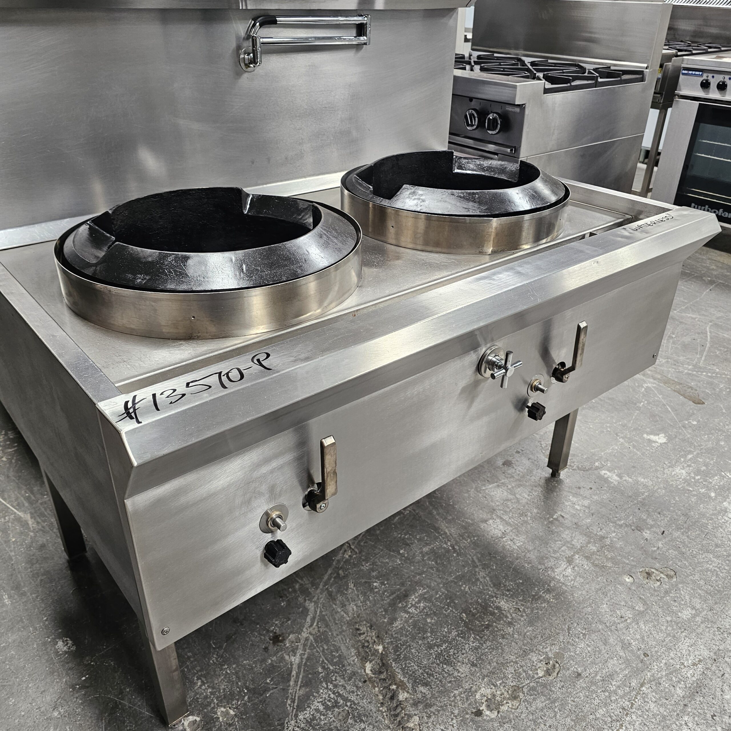 A stainless steel commercial wok range with two large circular burners, control knobs, and labeled #12570-P, situated in a professional kitchen setting on a concrete floor. MRCE