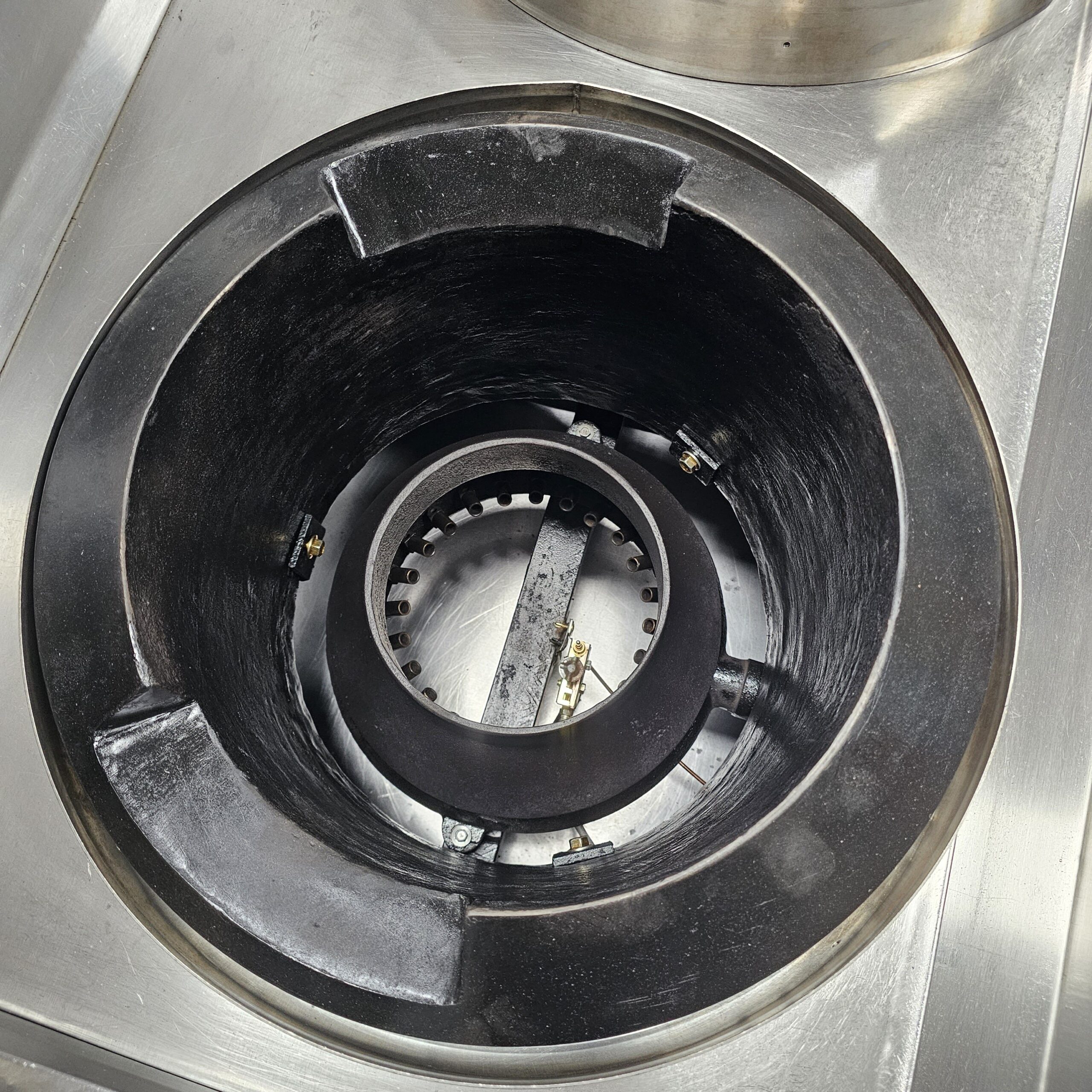 A close-up of a commercial gas stove burner with a circular metal grate and inner ring, set in a stainless steel countertop. The burner is clean and unused, showing gas outlets around the ring. MRCE