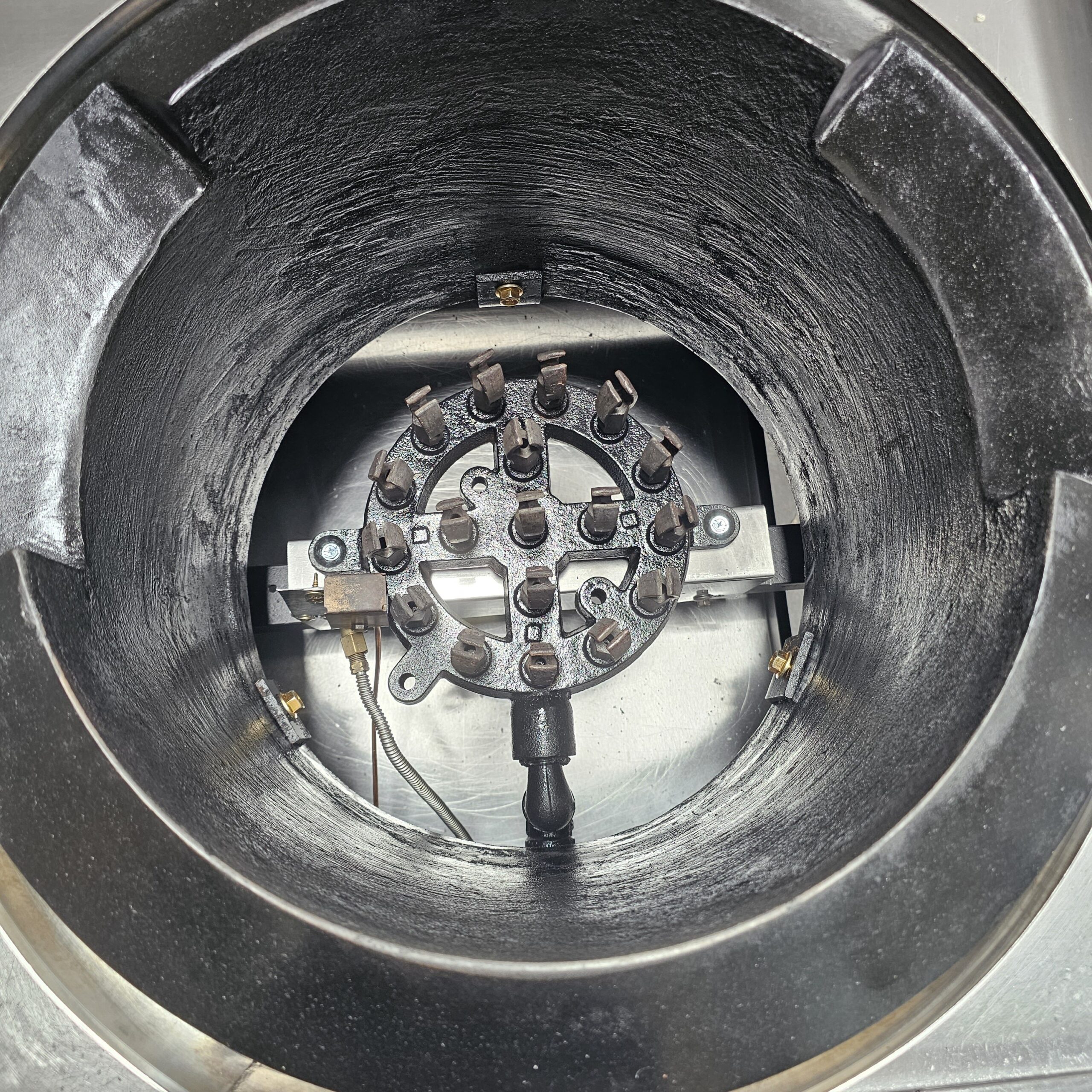 Close-up view of the inside of a commercial gas stove burner, showing the circular metal burner head with multiple gas outlets and igniter, surrounded by a black metal chamber. MRCE