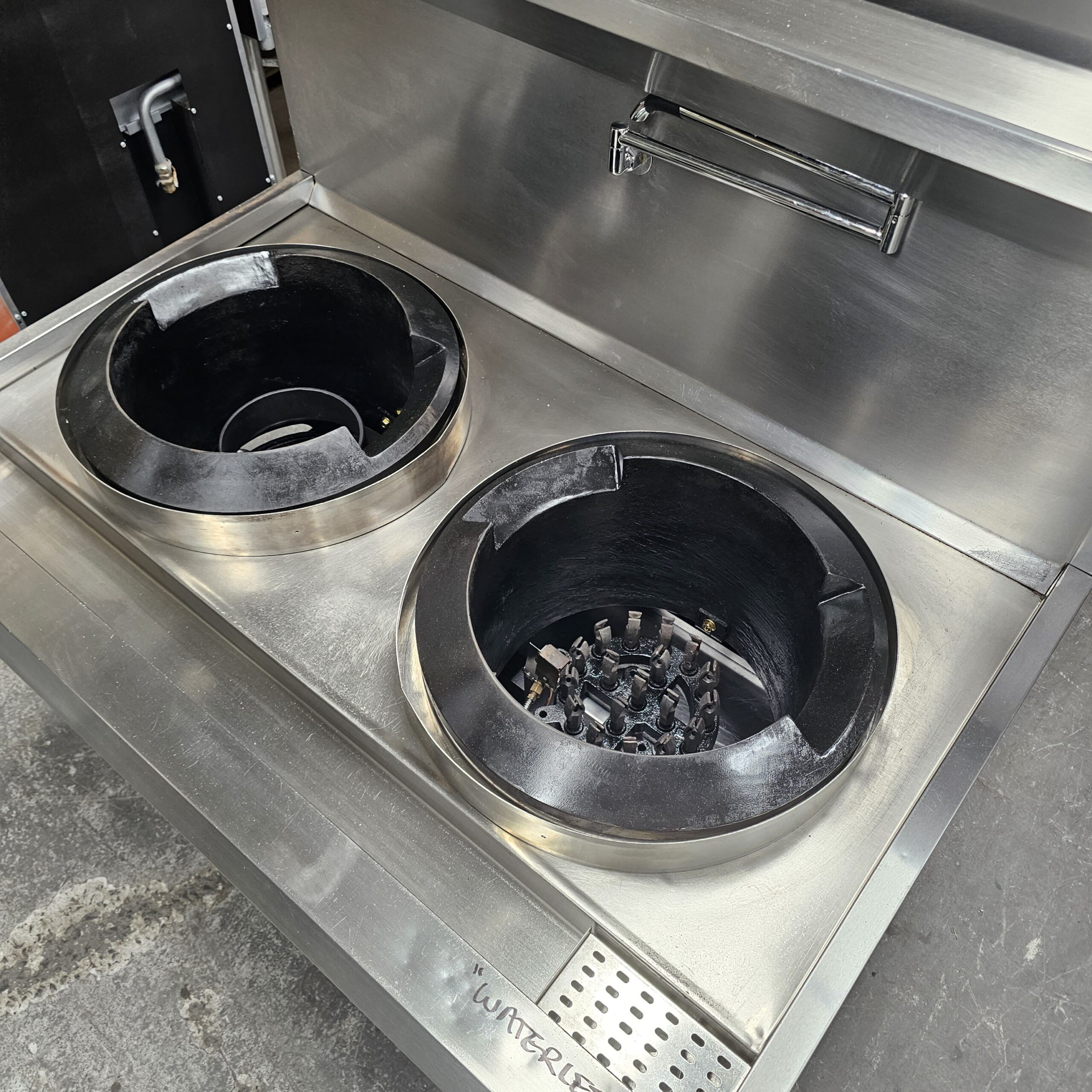 A stainless steel commercial kitchen stove with two large round burners, one featuring a gas flame ring and the other empty, set in a countertop with a small vented section labeled WATERFILL. MRCE
