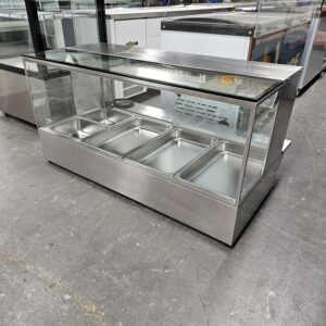 A stainless steel and glass refrigerated display case with five empty metal food trays inside, set on a concrete floor in a commercial or industrial environment. MRCE
