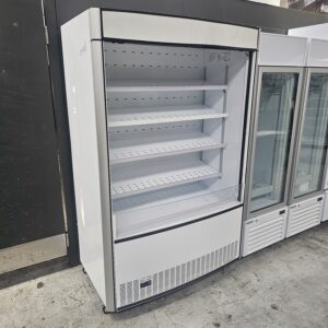 A large, empty, white refrigerated display case with four shelves is shown standing on a concrete floor next to other refrigeration units in a commercial setting. MRCE