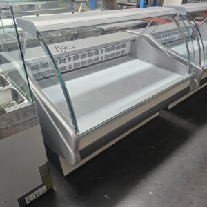 A large, empty refrigerated display case with curved glass and a white interior is positioned indoors on a dark floor, surrounded by other commercial kitchen equipment. A price is written on the glass in the background. MRCE