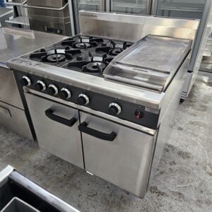 A stainless steel commercial kitchen stove with four gas burners, control knobs, a flat griddle, and two oven doors, situated in a professional kitchen setting. MRCE