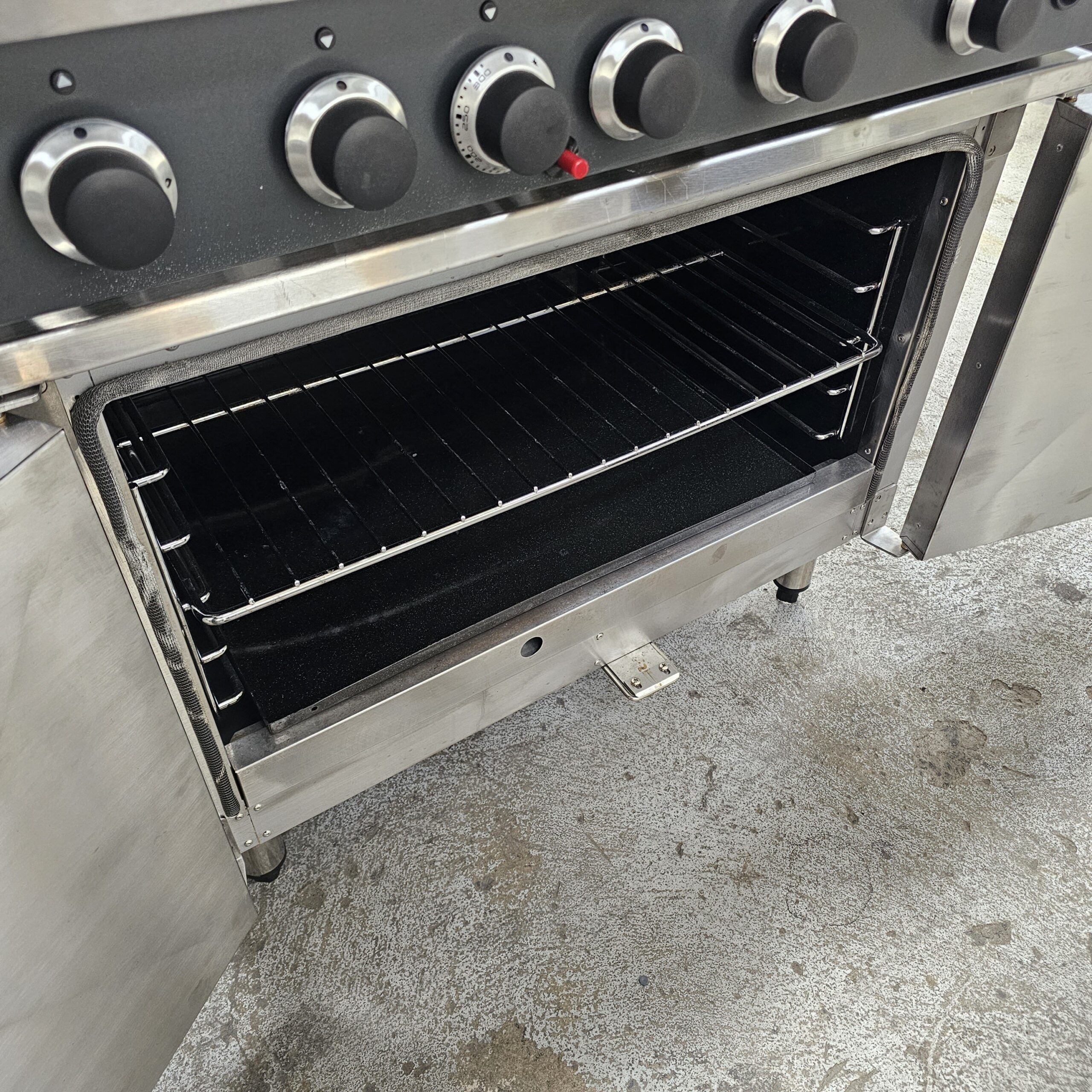 A stainless steel oven with its double doors open, showing two wire racks inside. The stovetop knobs and control panel are visible above the oven. The oven sits on a textured, gray floor. MRCE