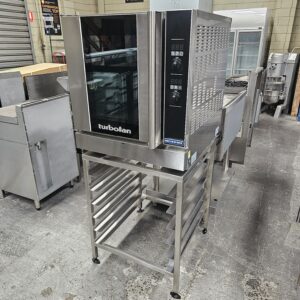 A commercial turbofan oven stands on a stainless steel rack with tray slots, surrounded by other industrial kitchen equipment in a commercial kitchen or equipment showroom. MRCE